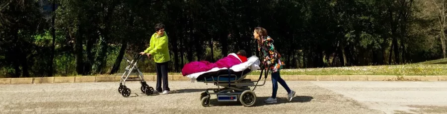 Dues persones passejant per un passeig, una amb el suport d'una caminador, i l'altra estirada en una cadira-llit que empeny una tercera persona